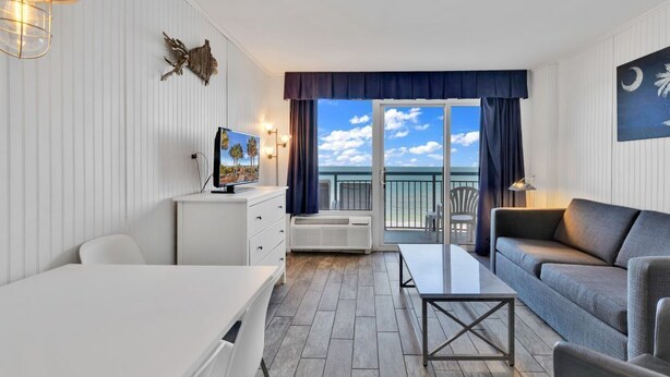 A modern living room with a sofa, a coffee table, and a TV on a white cabinet. There is a balcony with a view of the sea and a painting of a crescent moon on the wall. - Boardwalk Resort Condo Ocean-front Balcony 🏊 Pool 🏊 (Myrtle Beach)