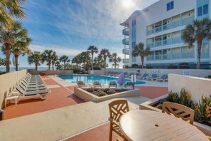 Un área de piscina de hotel con sillones de descanso y una mesa, rodeada de palmeras y un edificio con balcones.