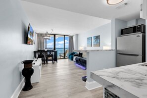 A modern kitchen with a large refrigerator, a dining area with a table and chairs, and a view of the ocean through large windows. - Modern Beachfront Condo Hosteeva 🏊 Pool 🏊 (Myrtle Beach)