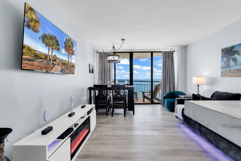 A modern hotel room with a balcony, a dining area, and a large TV screen displaying a beach scene.