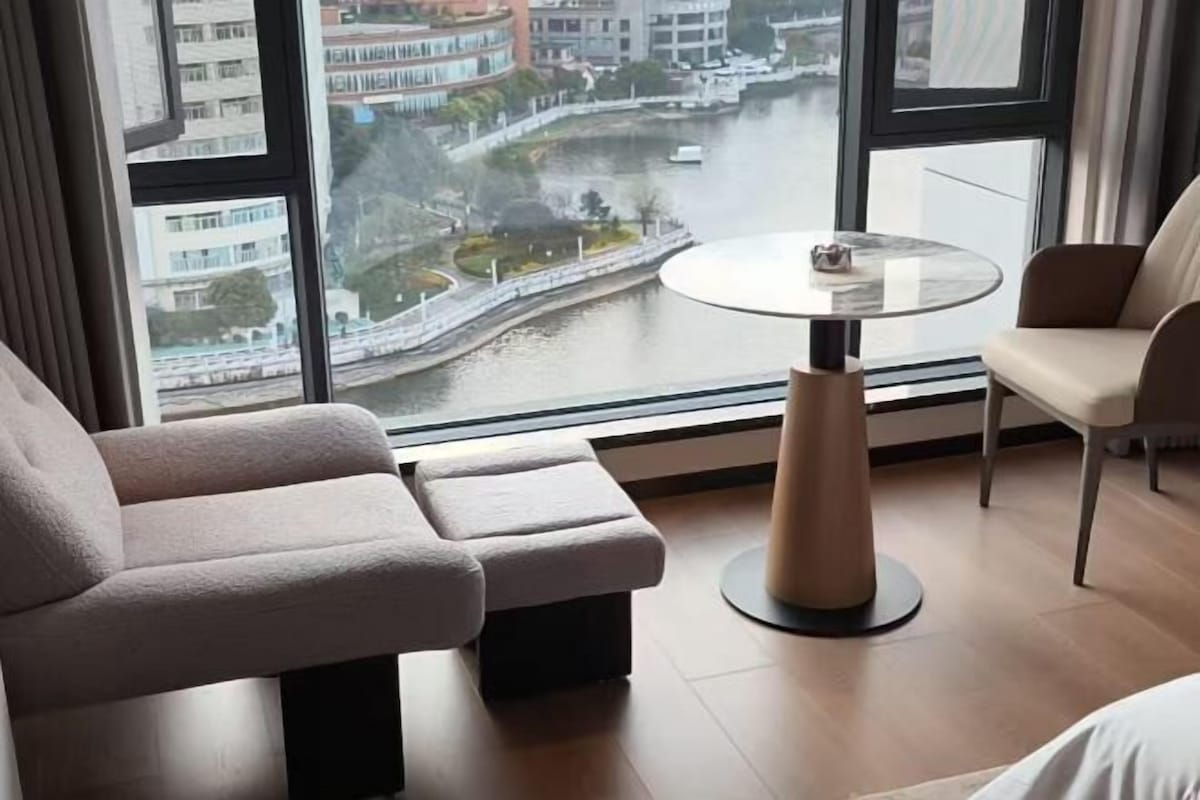 A modern hotel room with a large window offering a view of a river and buildings. There is a sofa, a small table, and a chair.