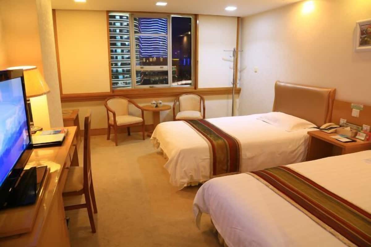 A hotel room with two beds, a desk with a computer, a chair, and a window with a view of buildings.