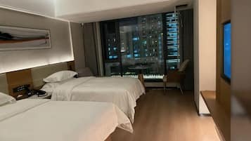 A hotel room with two beds, a desk, a chair, a TV, and a large window with a city view.
