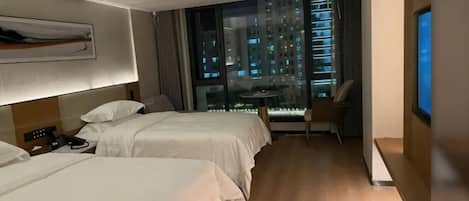 Habitación de hotel con dos camas, un escritorio, una silla, un televisor y un amplio ventanal con vista a la ciudad.