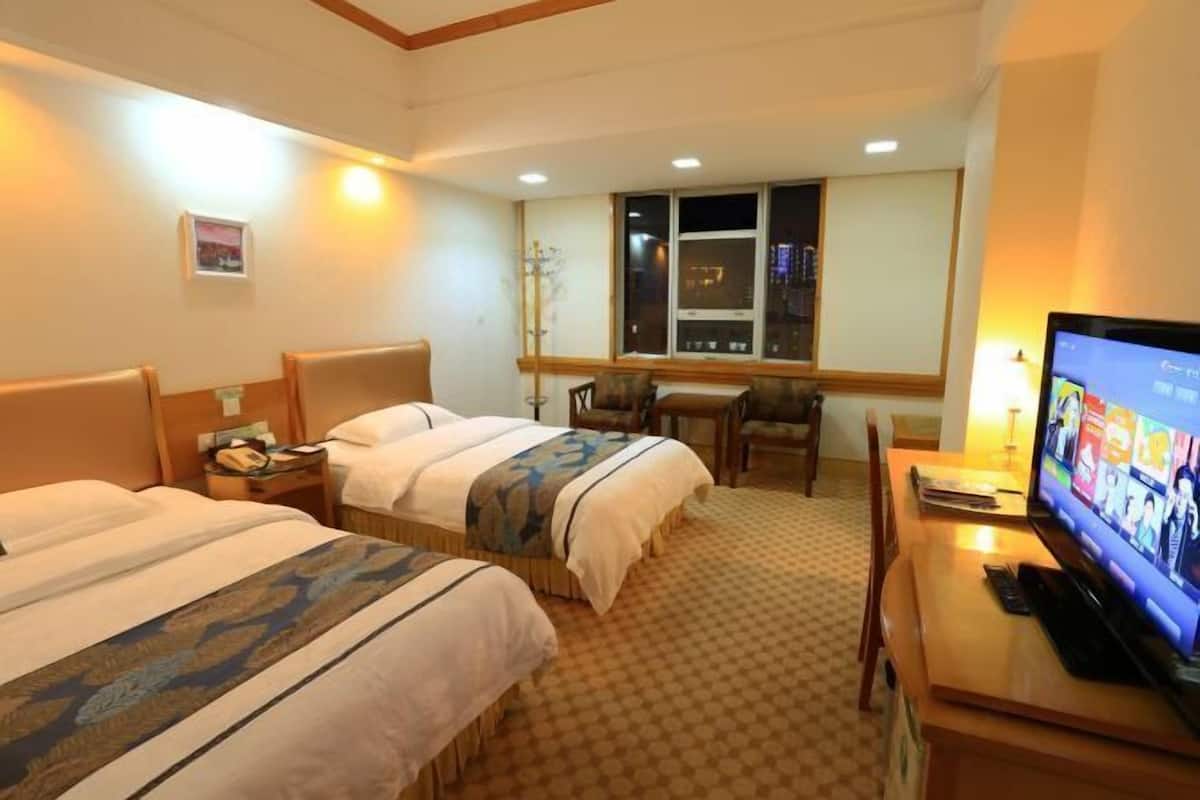 A hotel room with two beds, a television, a desk, and a window with a city view.