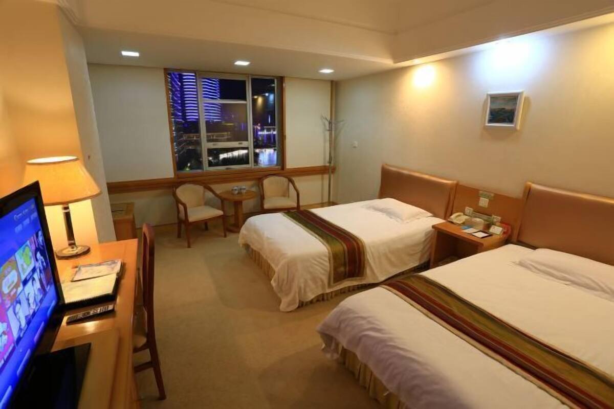 A hotel room with two beds, a desk with a computer, a chair, a small table, and a window with a view of a cityscape at night.