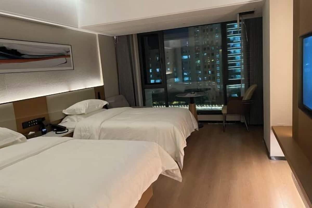 A hotel room with two beds, a desk, a chair, a TV, and a view of a cityscape through a large window.