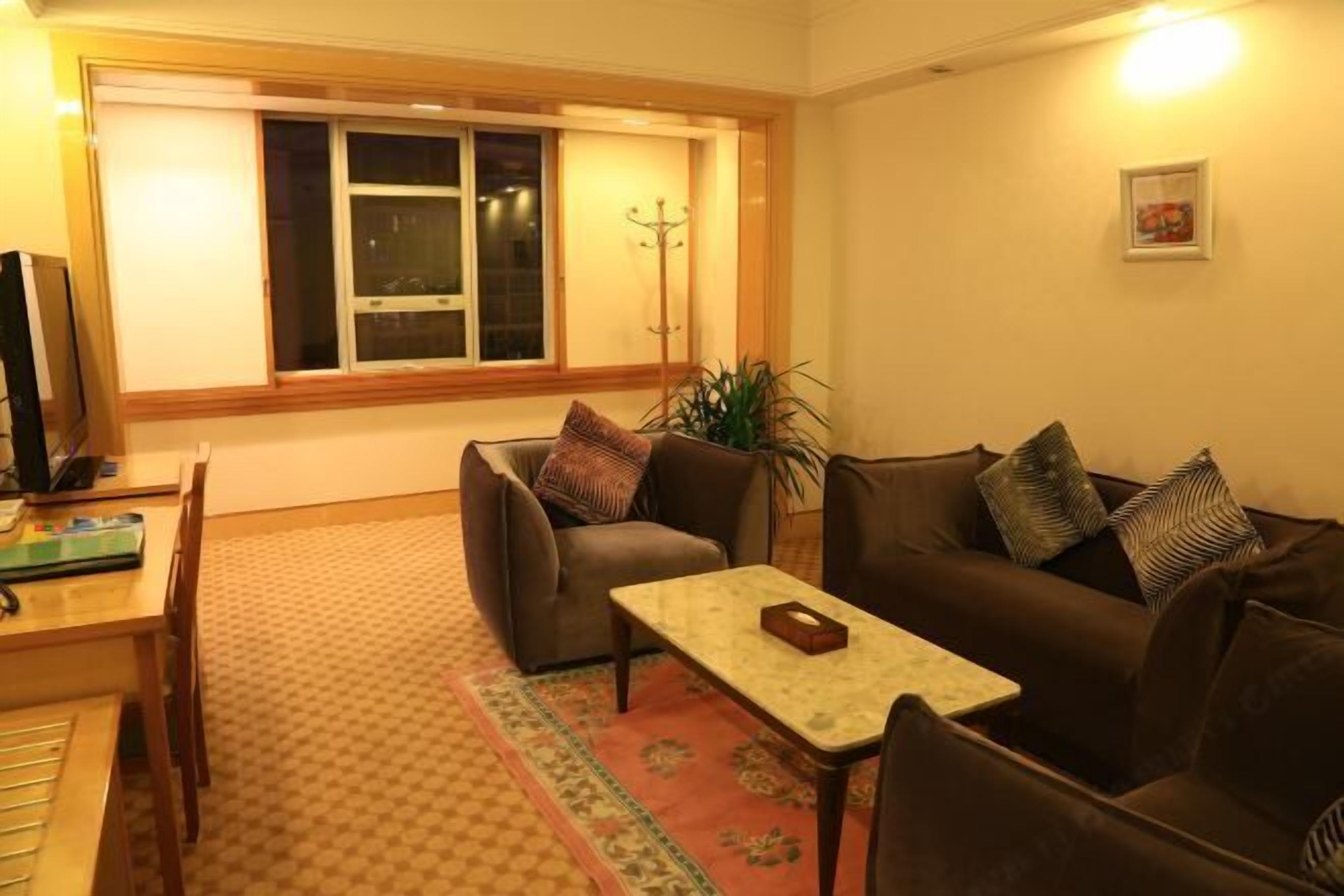 A living room with a sofa, a coffee table, and a TV on a wooden table. There is a window with blinds, a plant, and a coat rack.