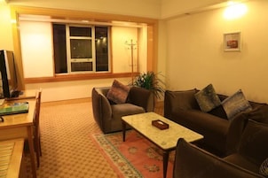 Sala de estar con sofá, una mesa de centro y un televisor sobre una mesa de madera. Se ve una ventana con persianas, una planta y un perchero.