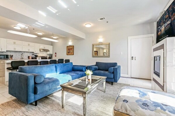 A modern living room with a blue sectional sofa, a glass coffee table, and a kitchen area with white cabinets and a microwave.