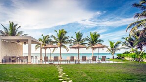 Zona junto a la piscina con sillones de descanso y sombrillas, palmeras y una vista despejada del océano.