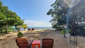 Un espace de détente aménagé sur la plage, avec deux chaises en bois et une petite table, entouré d’arbres et offrant une vue sur l’océan.
