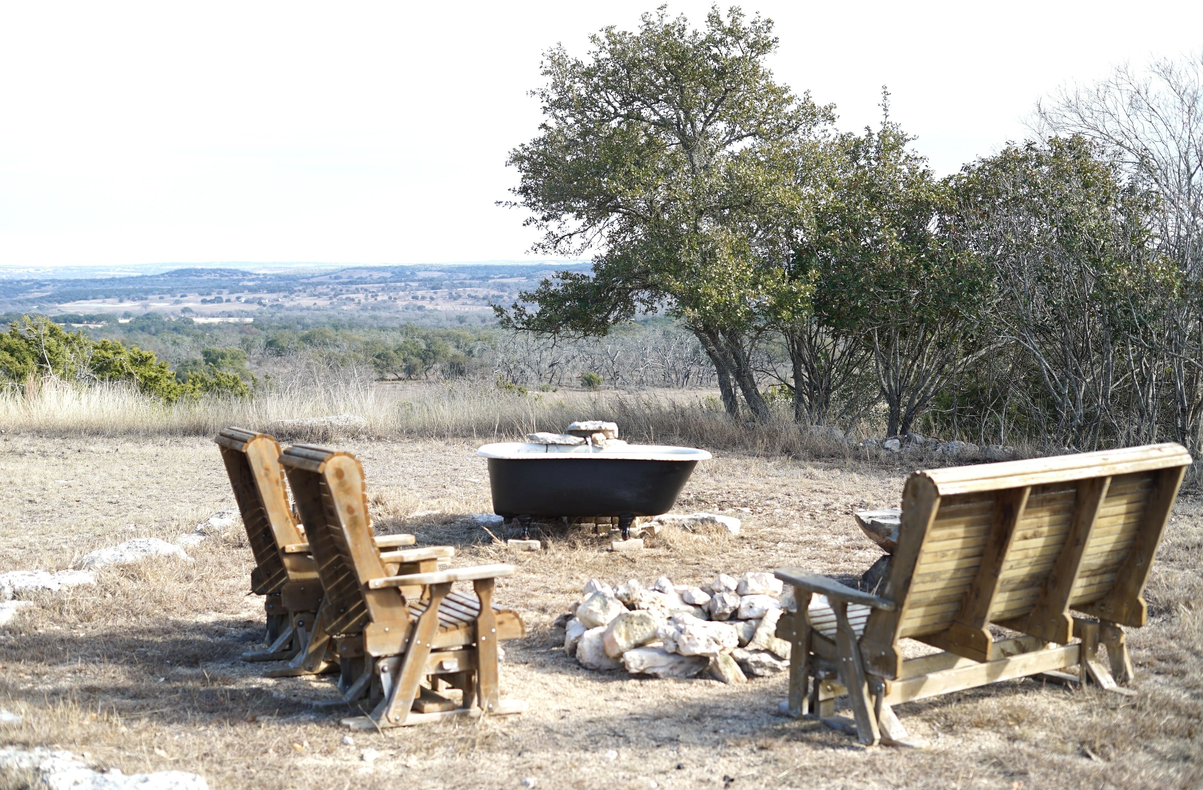 A rustic outdoor seating area with wooden chairs and a black bathtub, surrounded by dry grass and trees, overlooking a distant landscape.