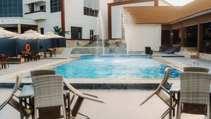 Une piscine avec une fontaine, entourée de fauteuils en osier et de parasols, avec un bâtiment en arrière-plan.
