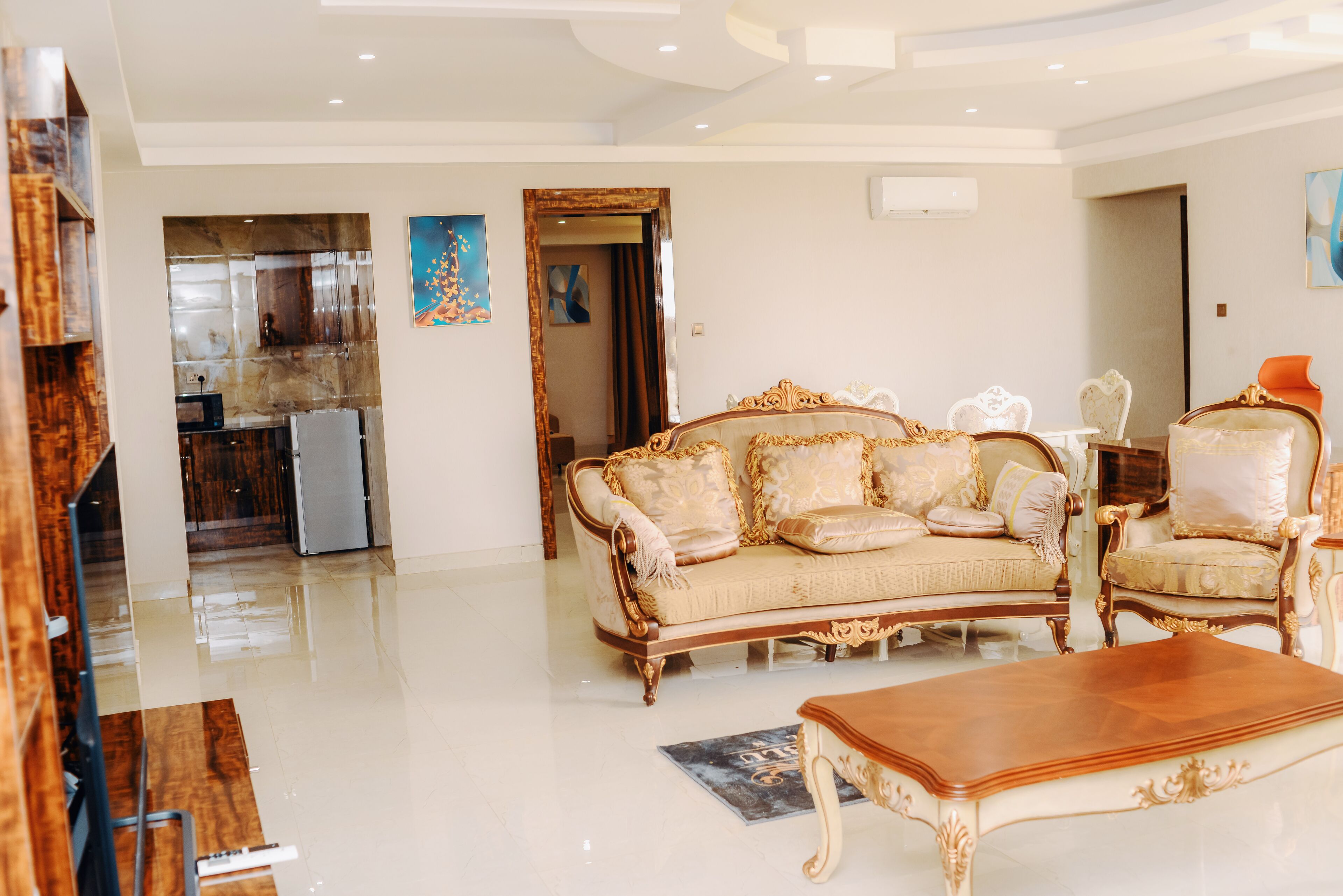 A spacious living area with a large ornate sofa, a wooden coffee table, and a matching armchair. A kitchen is visible in the background.