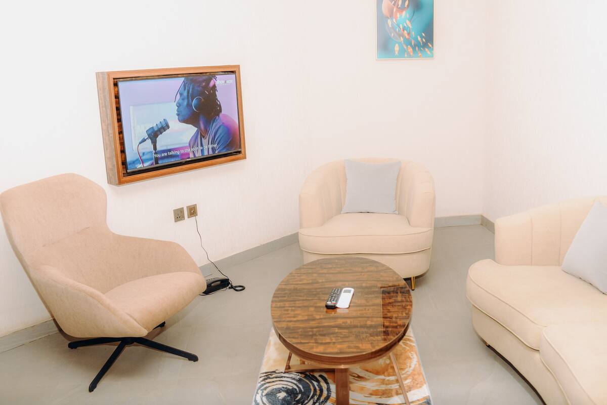 Ein Wohnzimmer mit einem beigefarbenen Sofa, einem beigefarbenen Sessel und einem runden Couchtisch mit einer Fernbedienung. Ein an der Wand befestigtes Fernsehgerät zeigt eine Person, die in ein Mikrofon spricht.