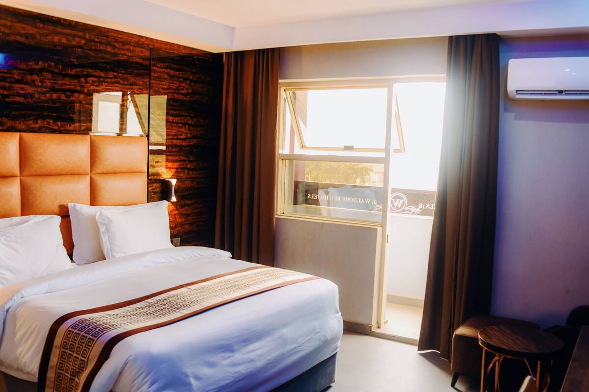 Ein Hotelzimmer mit einem großen Bett, einem hölzernen Kopfteil, einem Fenster mit Vorhängen und einer Klimaanlage an der Wand.