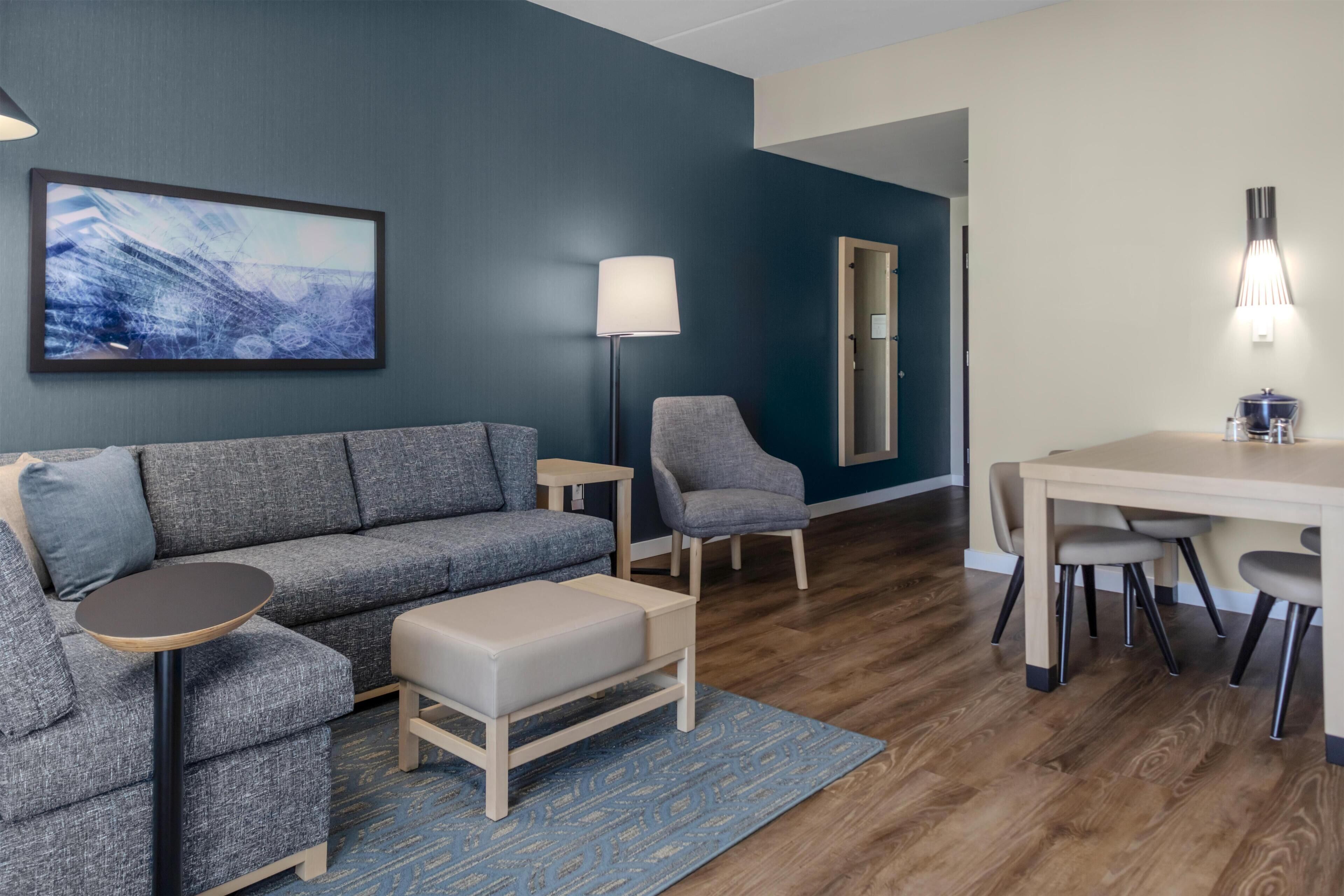 A modern living room with a grey sofa, a small round table, a grey armchair, a wooden coffee table, and a dining area with a table and chairs.