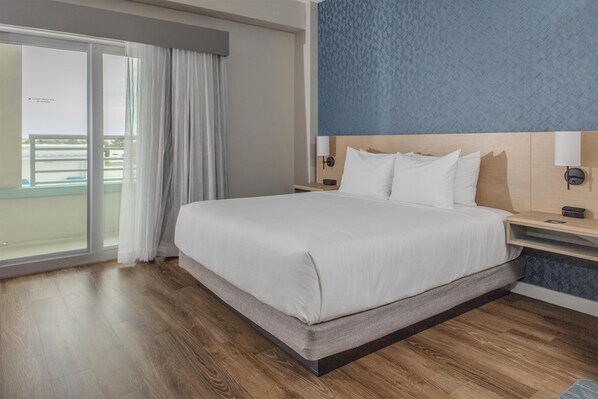Habitación de hotel con una cama grande, una mesita de noche, una lámpara de pared y vistas al balcón.