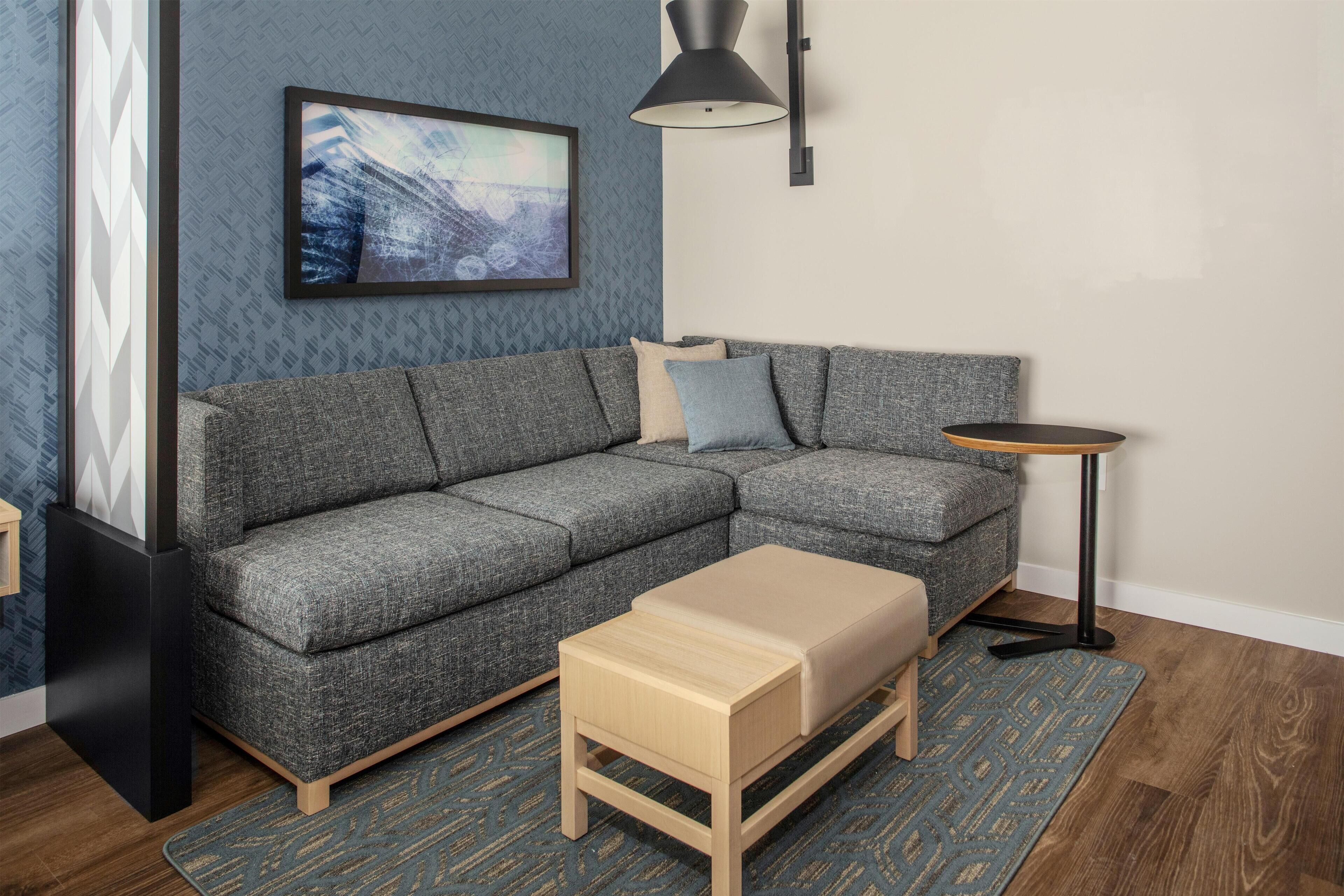 A modern living room with a grey sectional sofa, a small wooden coffee table, a blue patterned rug, a black floor lamp, and a framed picture on the wall.
