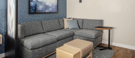 A modern living room with a grey sectional sofa, a small wooden coffee table, a blue patterned rug, a black floor lamp, and a framed picture on the wall.