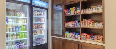 A kitchenette with a refrigerator stocked with beverages and snacks, and wooden cabinets above holding various food items.