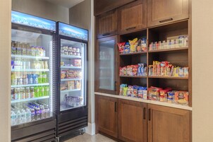 A kitchenette with a refrigerator stocked with beverages and snacks, and wooden cabinets above holding various food items.