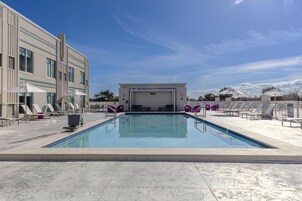 A swimming pool with lounge chairs and umbrellas, a covered seating area, and a building with large windows.