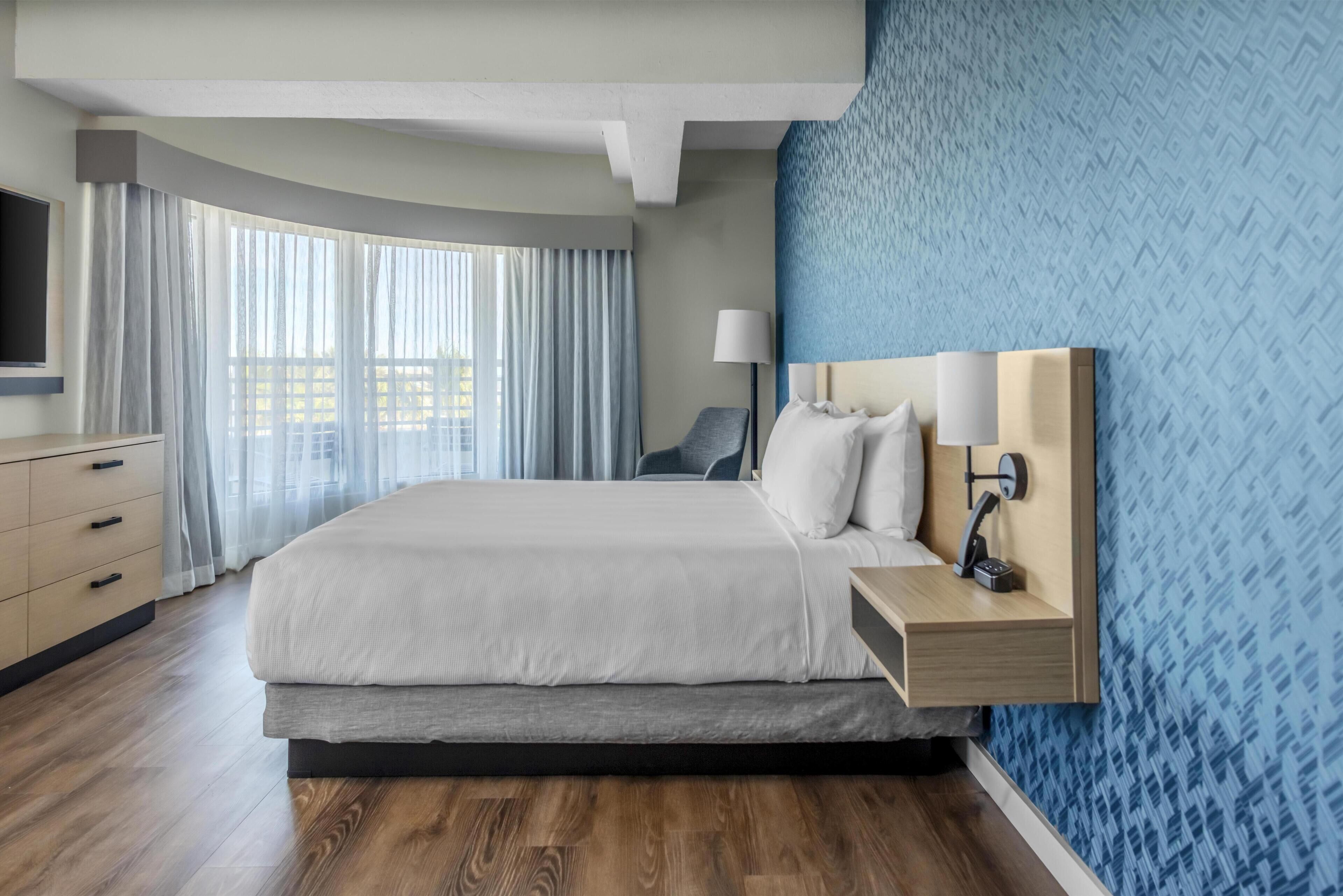A hotel room with a large bed, a bedside table with lamps, a TV, a desk with drawers, and a window with curtains.