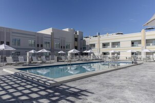 Uma área de piscina com espreguiçadeiras e guarda-sóis, rodeada por um edifício moderno com varandas.