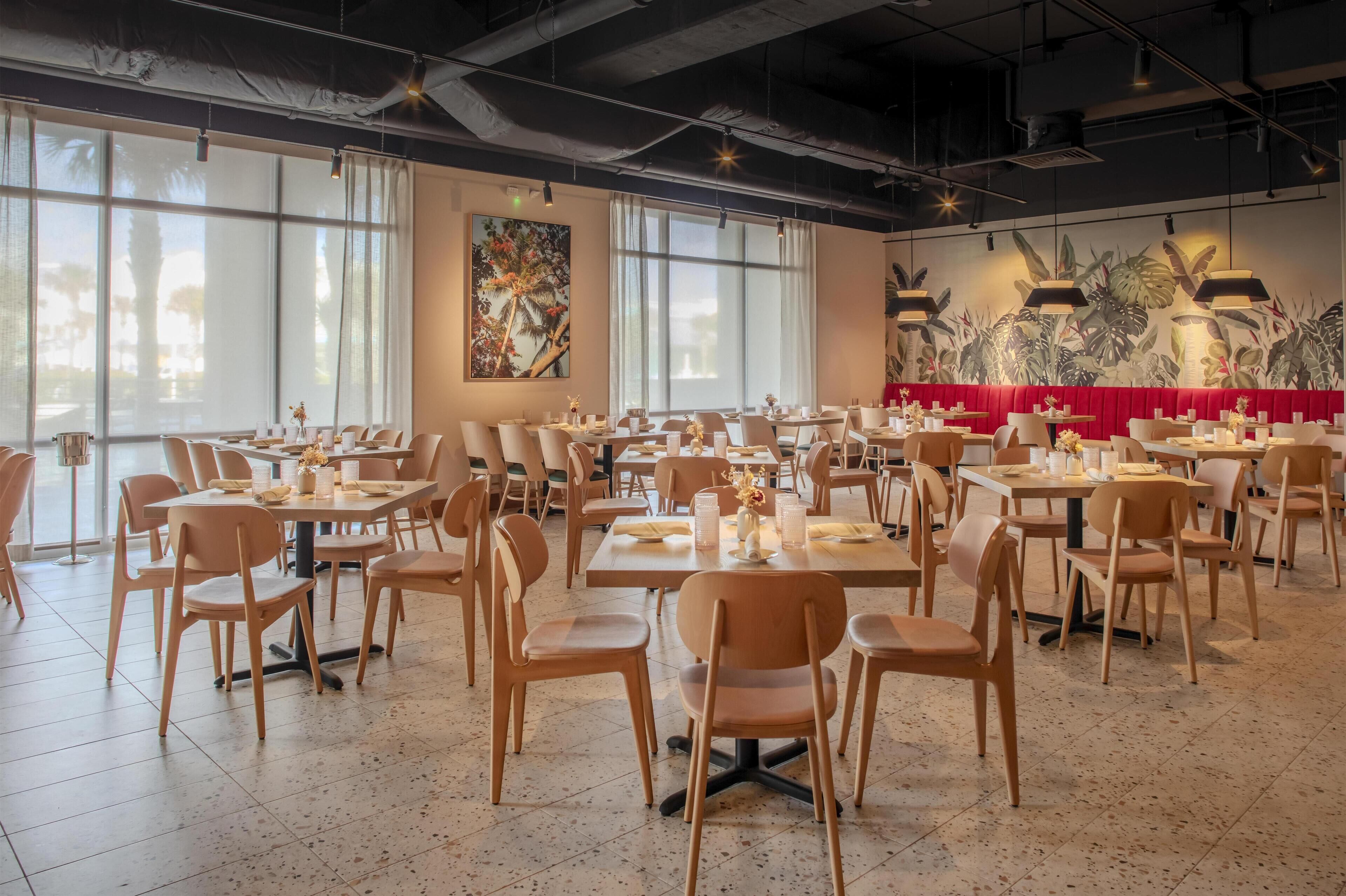 Un restaurant spacieux avec de hauts plafonds, de grandes fenêtres et un coin salon avec des banquettes rouges. Les tables sont disposées de manière ordonnée, accompagnées de chaises, et le sol est carrelé.