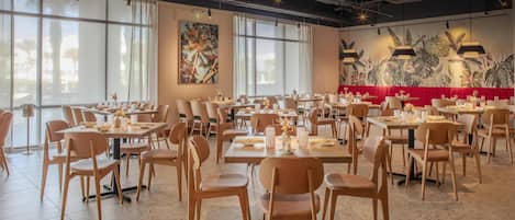 A spacious restaurant with high ceilings, large windows, and a red booth area. Tables are neatly arranged with chairs, and the floor is tiled.