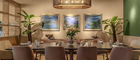A dining area with a large table, cushioned chairs, a central vase with flowers, and framed artwork on the wall.