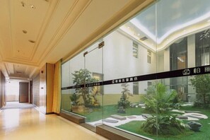 A hotel corridor with a glass wall, a reception desk, and a view of a courtyard with plants.
