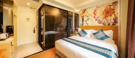 A hotel room with a bed, a desk, a chair, a door, and a large floral artwork on the wall.