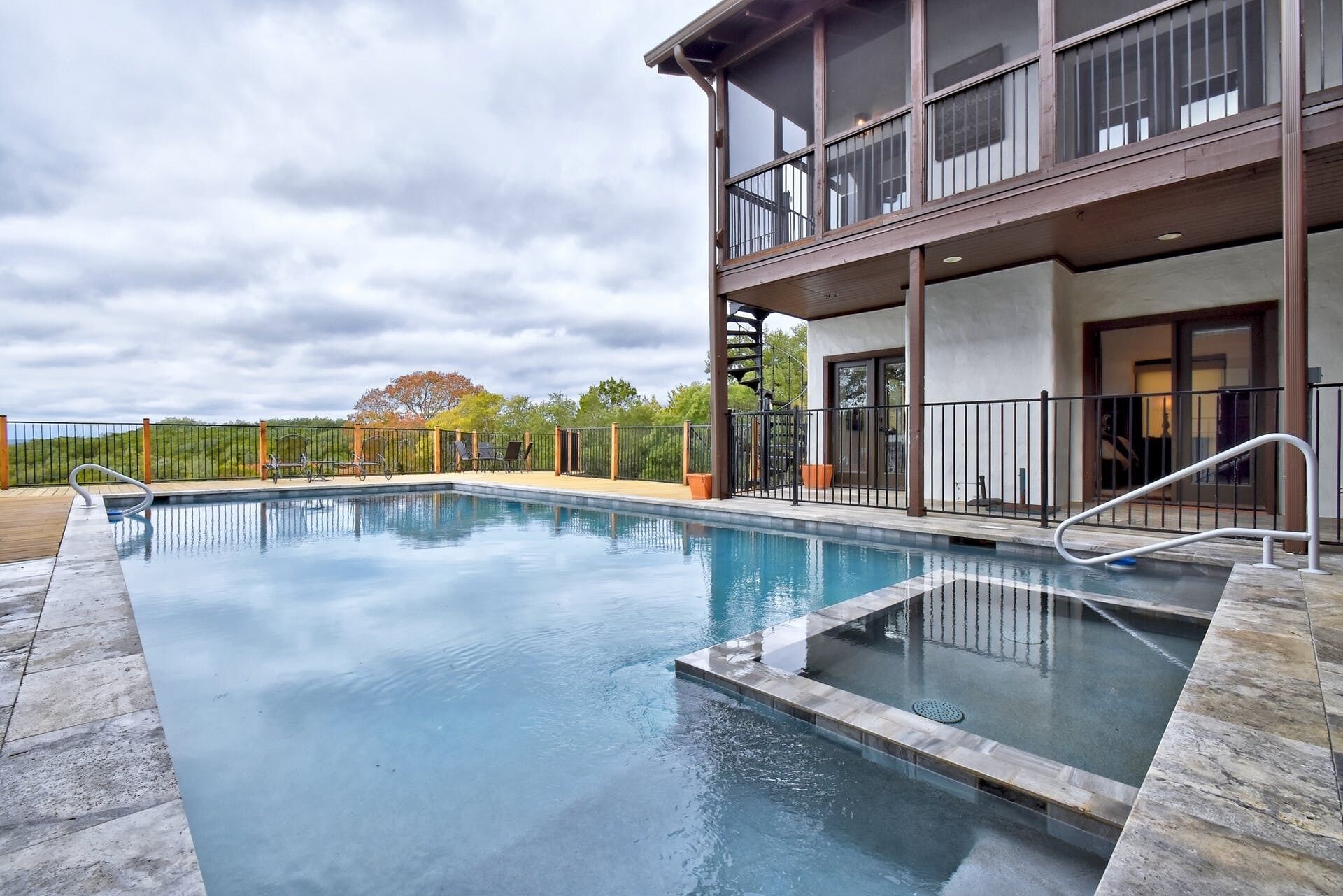 A rectangular swimming pool with a wooden deck surrounding it, a covered seating area with chairs, and a building with a balcony and large windows.