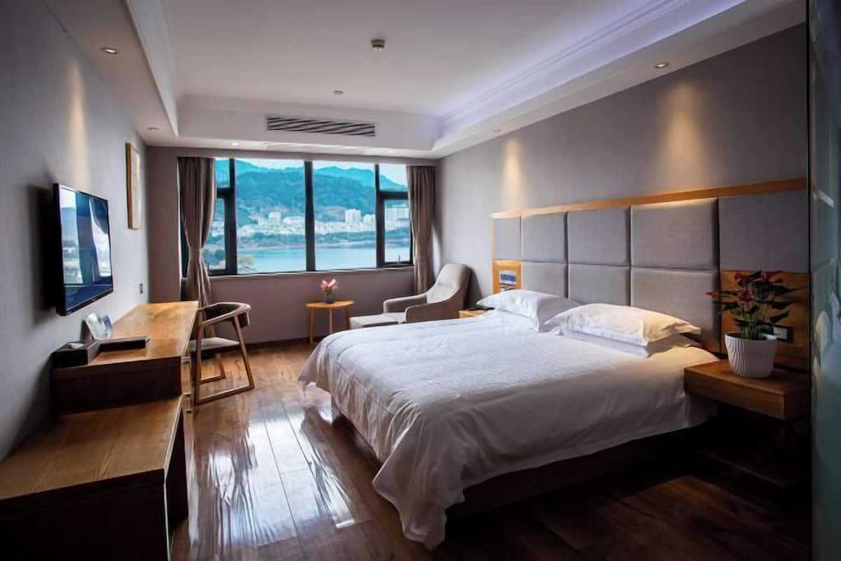 A modern hotel room with a large bed, a desk, a chair, a TV, and a view of a city and mountains.