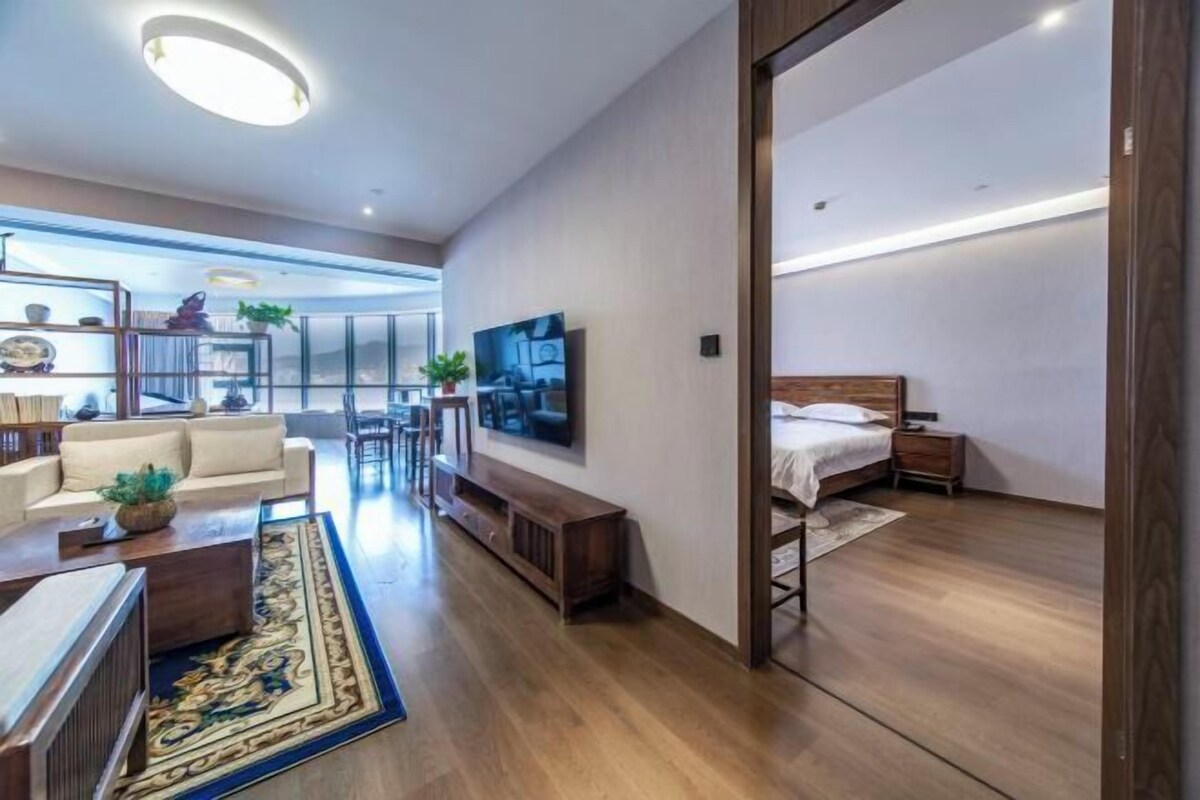 A modern living room with a sofa, coffee table, and TV. There's a wooden floor and a bed in the adjacent room.