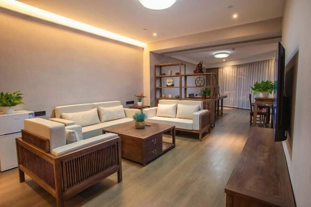 A modern living room with a sofa set, coffee table, and wooden side table. There is a television mounted on the wall and a small refrigerator in the corner.