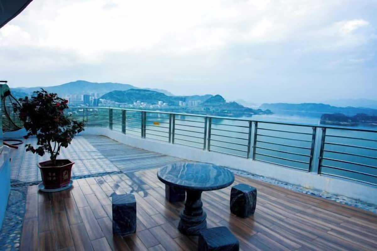 A wooden deck with a round table and potted plants, overlooking a cityscape and mountains.