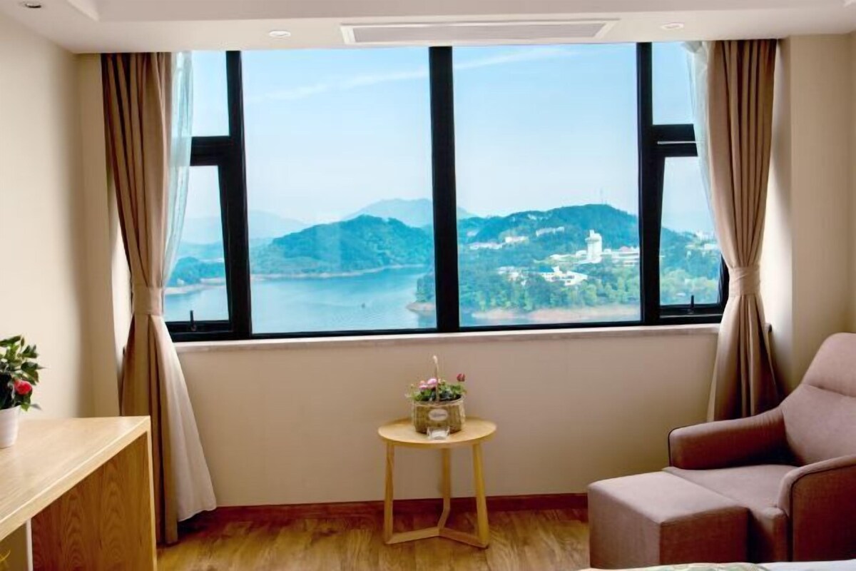 A room with a large window offering a scenic view of a lake and hills. There is a small round table with a plant and a chair.