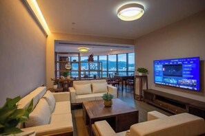 A modern living room with a large flat-screen TV, comfortable seating, a coffee table, and a view of the city through large windows.