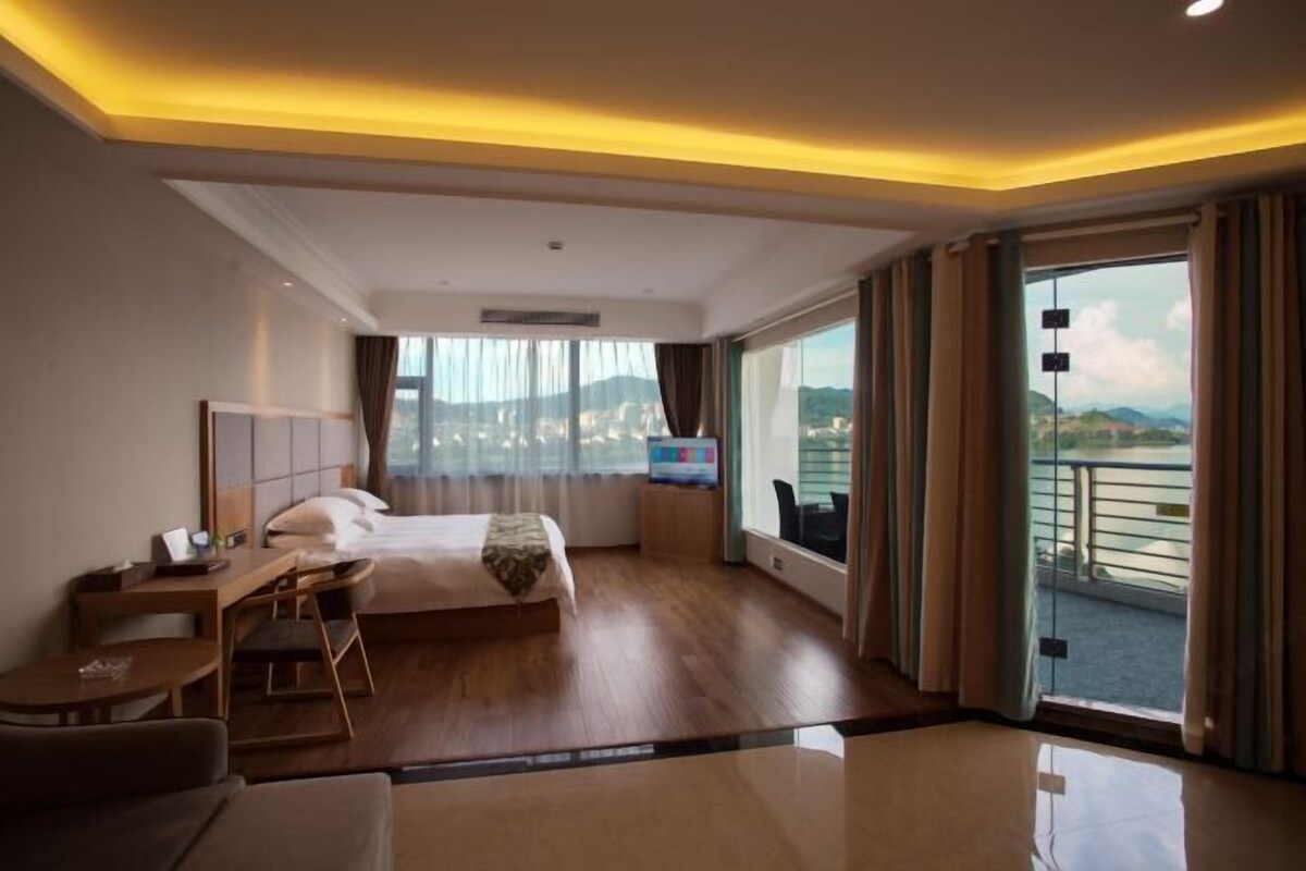 A hotel room with a bed, a desk, a chair, a TV, and a balcony with a view of the city.