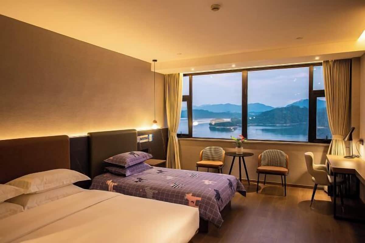 A hotel room with two beds, a desk, chairs, and a large window with a view of mountains and a body of water.