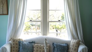 Une pièce avec une fenêtre, un canapé blanc orné de coussins à motifs floraux et de rideaux légers.