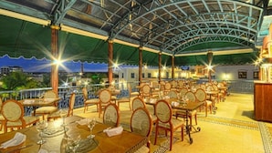 An outdoor dining area with tables and chairs, a covered roof, and a view of the cityscape.