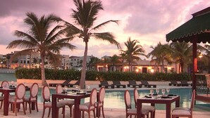 A resort pool area with tables and chairs, palm trees, and buildings in the background.