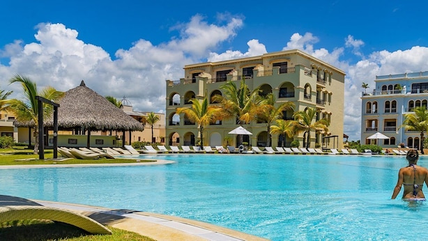 A resort pool area with a large pool, lounge chairs, palm trees, and a building with multiple balconies.
