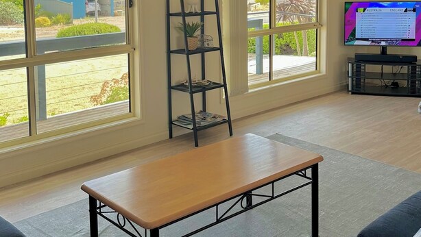 Un salon spacieux avec de grandes fenêtres, une table en bois, des canapés noirs, un téléviseur et une horloge murale.