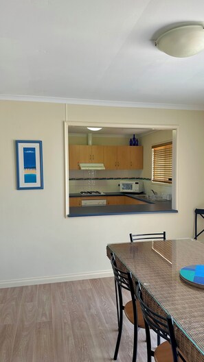 A dining area with a glass table and chairs, a kitchen visible through an archway, and a framed picture on the wall.